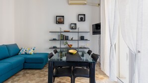 A modern living room with a blue sofa, a glass dining table with chairs, and a white wall with shelves containing books and decorative items.