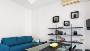 A modern living room with a blue sofa, a glass dining table, and a kitchen area with shelves and a microwave.