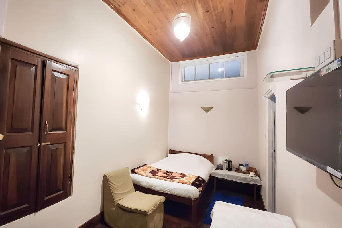 Um quarto pequeno e acolhedor com teto de madeira, uma cama com manta estampada, uma cadeira, uma mesinha com chaleira e uma TV fixada na parede.