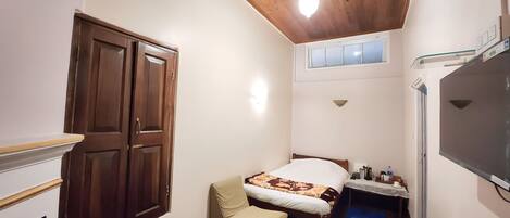 A small, cozy room with a wooden ceiling, a bed with a patterned blanket, a chair, a small table with a kettle, and a wall-mounted TV.