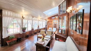 Una habitación amplia con piso de madera, ventanas grandes con cortinas blancas, un techo alto con tragaluces y un rincón de descanso acogedor con sofá y sillones.
