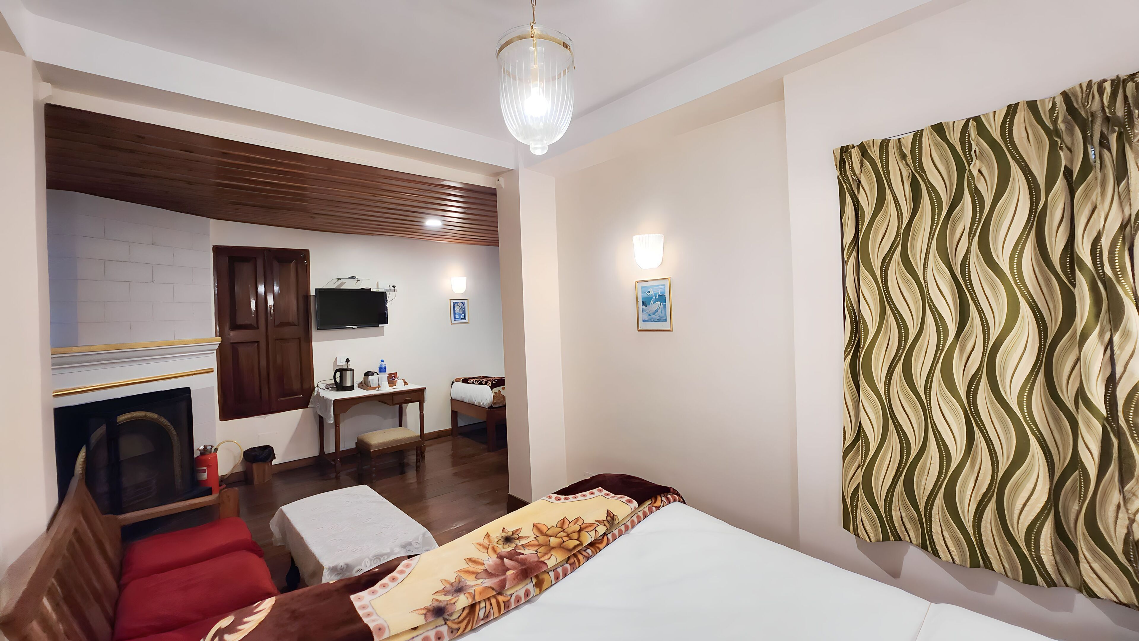 A hotel room with a bed, a wooden bench, a small table with a kettle, a television, and a patterned curtain.