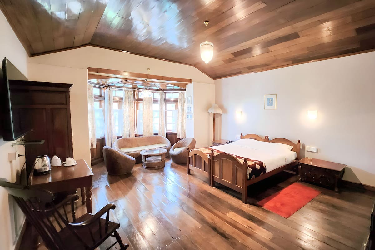 Quarto com teto de madeira, cama com cabeceira de madeira, uma cadeira, uma mesa e uma janela com cortinas.