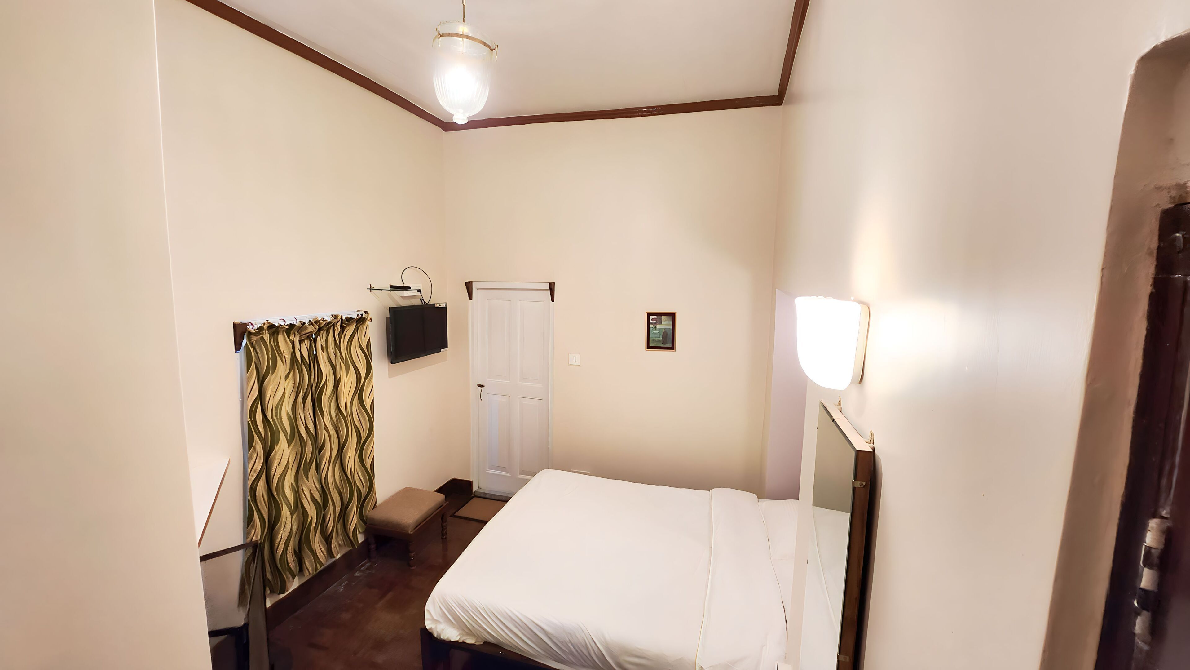 Een kleine, netjes ingerichte slaapkamer met een bed, nachtkastje, spiegel, deur en gordijn.