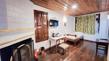 A room with a wooden ceiling, a bed, a desk with a chair, a fireplace, and a television.