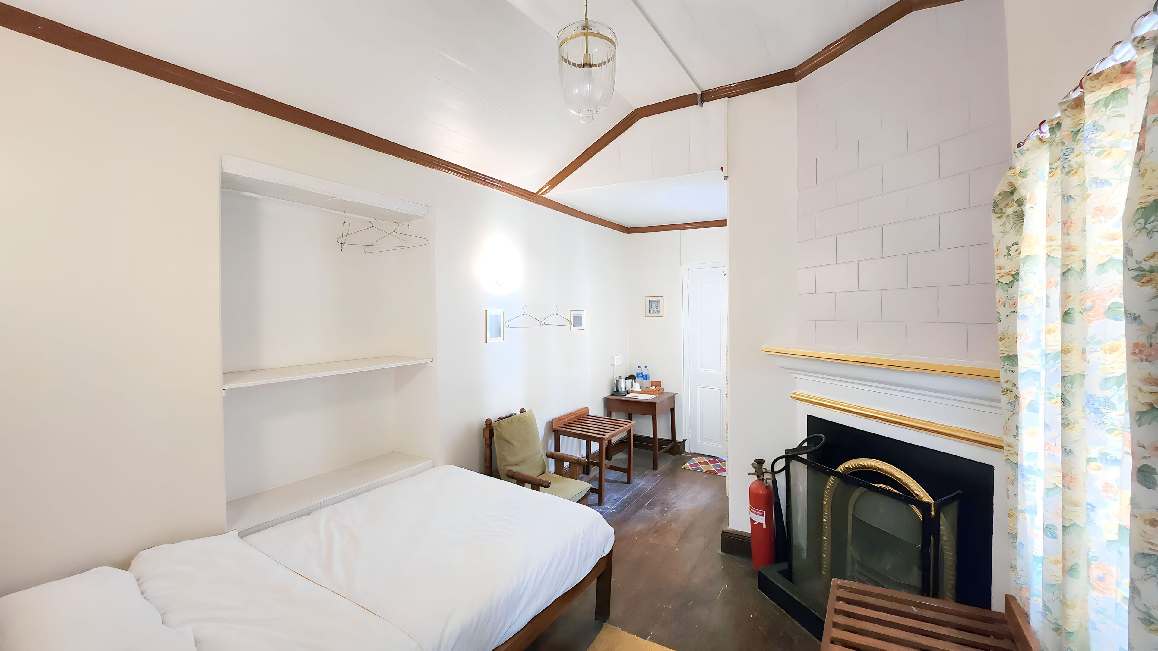 A bedroom with a bed, a wooden chair, a small table with a kettle, a fireplace, and a hanging light fixture.