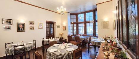 A dining area with checkered tablecloths, wooden chairs, a chandelier, and large windows.