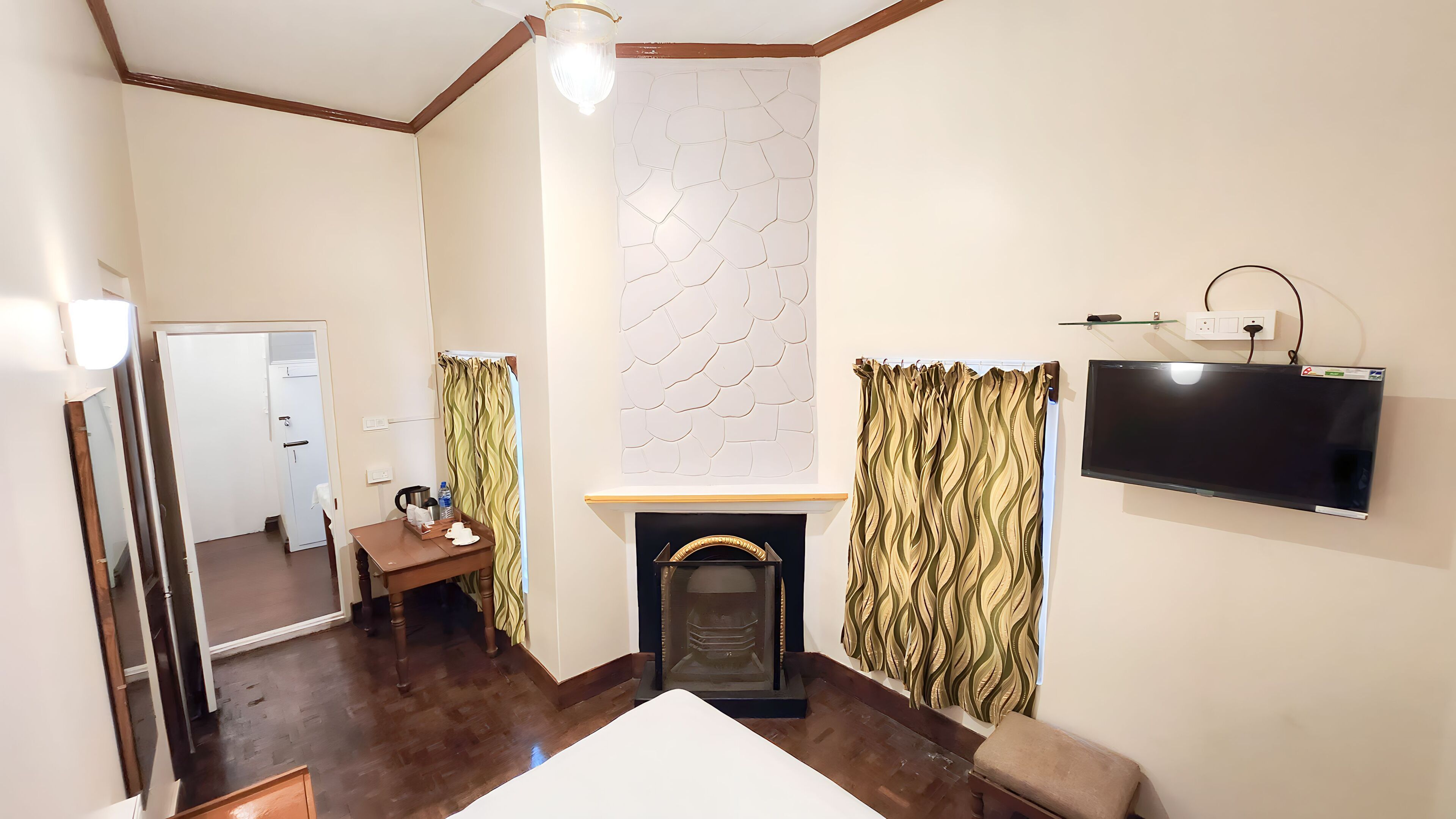 A hotel room with a fireplace, a flat-screen TV mounted on the wall, a wooden table with a tray, and a mirror.