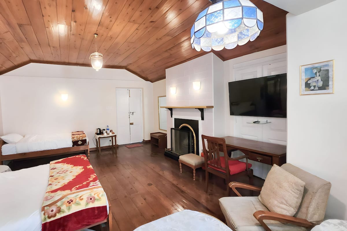 Quarto com teto de madeira, lareira, cama com colcha florida, sofá, poltrona e televisão na parede.