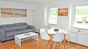 A modern living room with a grey sofa, white coffee table, and dining table with chairs. There are two windows with views of greenery and a parked car. - 1 Bedroom Apartment in Sellin (Sellin)