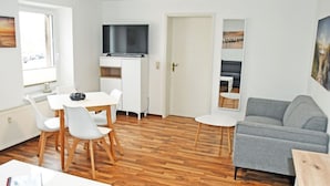 A modern living room with a grey sofa, a dining table with chairs, a flat-screen TV mounted above the TV stand, and a small side table. - 1 Bedroom Apartment in Sellin (Sellin)