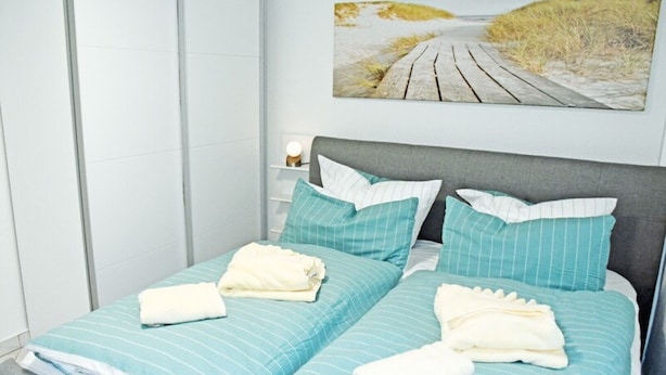 A double bed with blue-striped bedding and white pillows, a grey headboard, and a painting of a beach path on the wall. - 1 Bedroom Apartment in Sellin (Sellin)