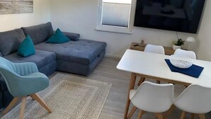 A modern living room with a grey sectional sofa, teal cushions, a white dining table with chairs, a flat-screen TV, and a small side table with a plant. - Vacation apartment 10 lake view (Sellin)