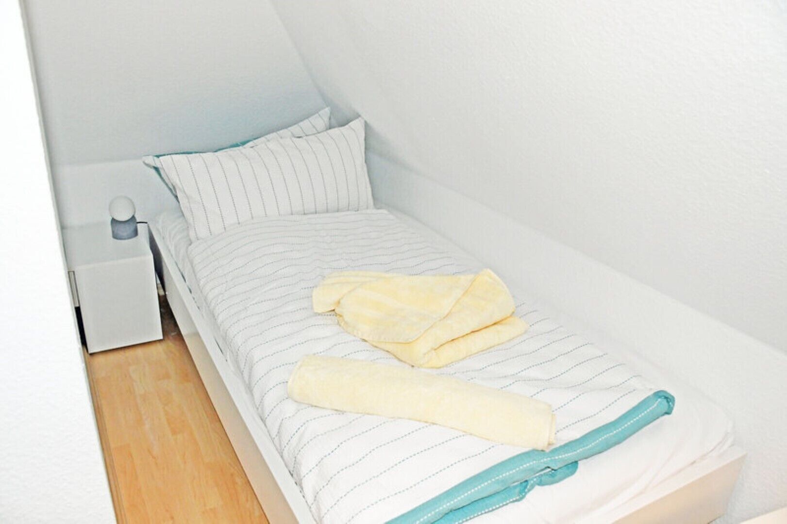 Una habitación pequeña con cama individual, colcha a rayas, almohada blanca y manta color turquesa.