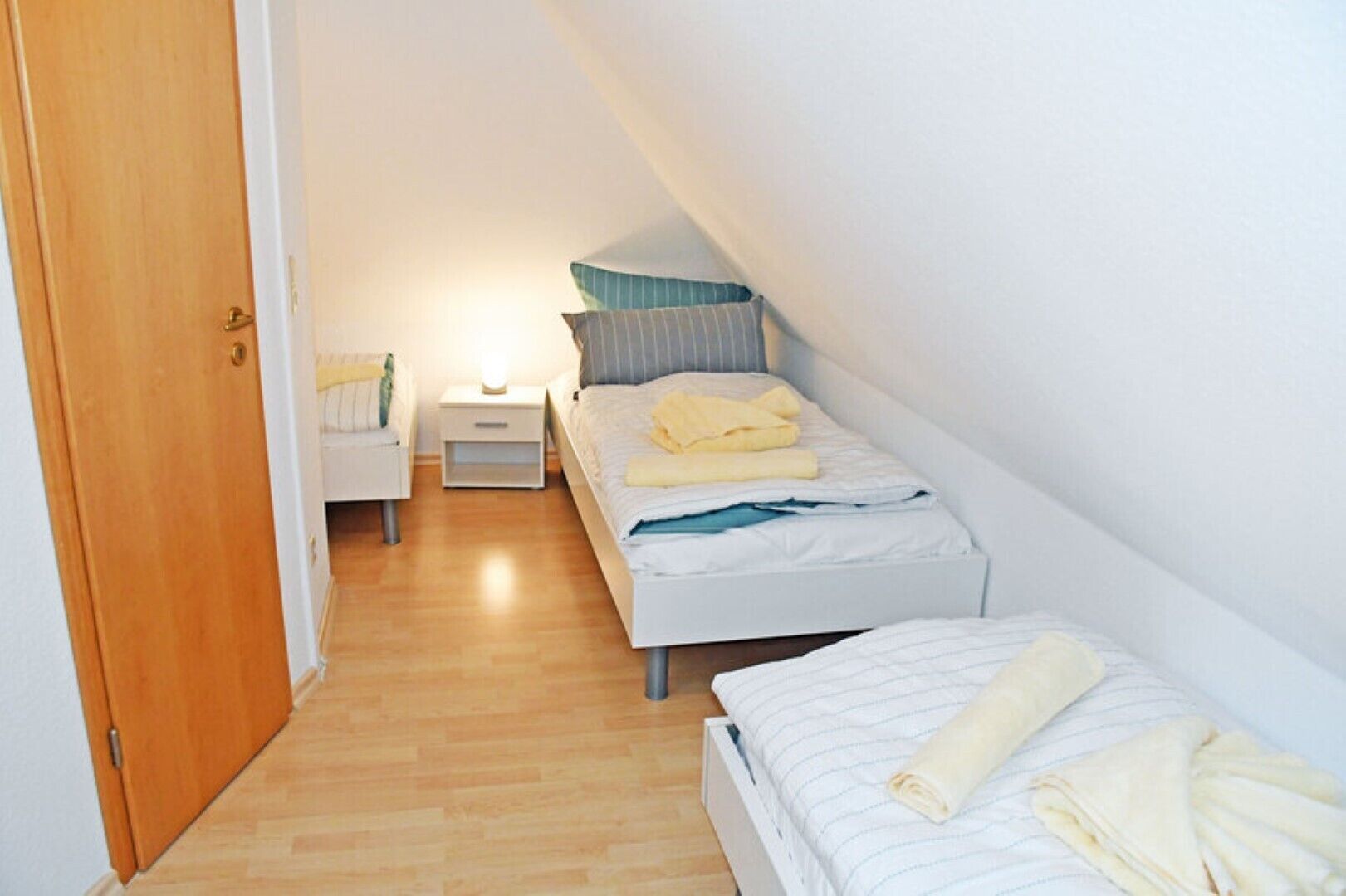 A small, well-lit room with a wooden floor, a single bed with a striped bedspread, a nightstand with a lamp, and a wooden door.