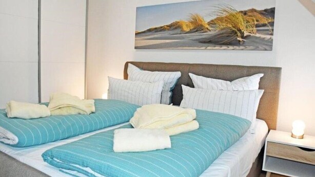 A neatly made bed with striped bedding and two pillows, a bedside table with a lamp, and a painting of a beach scene on the wall. - Ferienwohnung 6 Jasmund (Sellin)