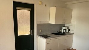 Una cocina compacta con una puerta oscura, una ventana, armarios blancos, un lavabo y una cafetera.