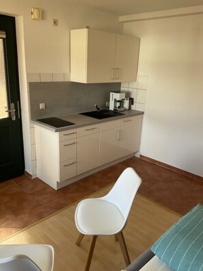 Une cuisine compacte comprenant un évier, un plan de travail et des placards. On y trouve une chaise blanche et une petite table avec un coussin vert.