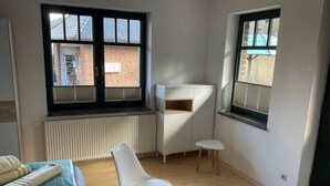 Una habitación pequeña y luminosa con un radiador, una silla blanca, una mesita y un sofá con una toalla doblada.