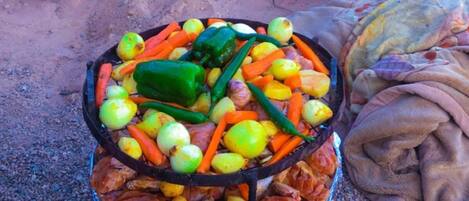 A grill with various vegetables and meat, a shovel, and a pot of sauce on a gravel surface.