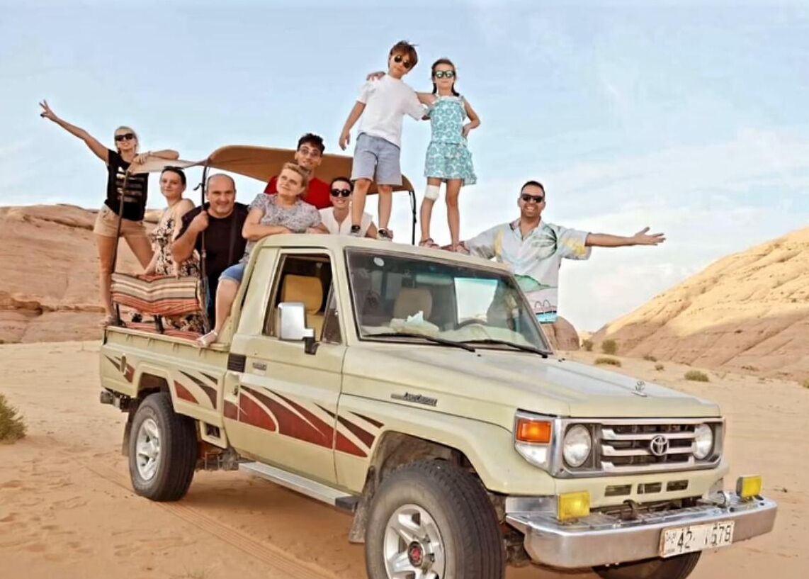 Een beige pick-up truck met een groep mensen die erop staan, in een woestijnlandschap.