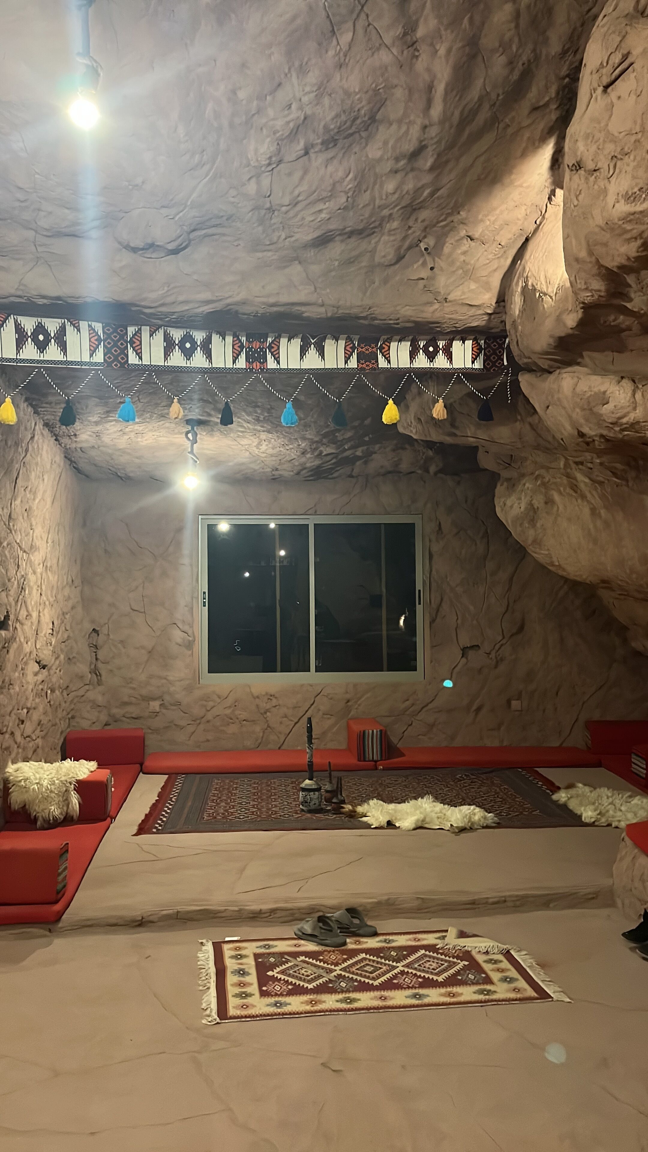 A cave-like room with a large window, red seating, a patterned rug, and hanging lights.