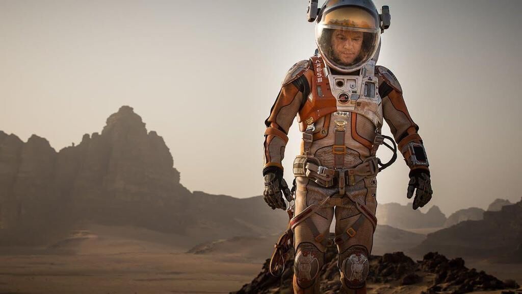 A person in a space suit standing on a rocky terrain with mountains in the background.
