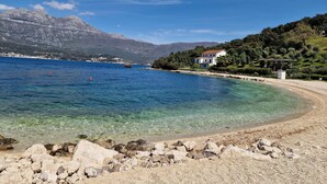 A coastal scene with a clear beach, rocky shoreline, and a house with a white facade and red roof. - Agape Rose (Klinci)