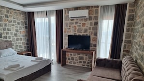 A hotel room with a stone wall, a bed with white linens, a flat-screen TV on a wooden stand, a brown sofa, and a window with curtains. - Agape Rose (Klinci)