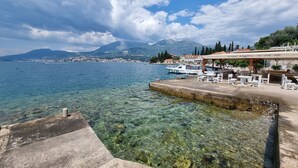 A waterfront restaurant with outdoor seating, clear water, and a mountain backdrop. - Agape Rose (Klinci)
