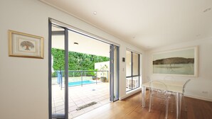 Una habitación amplia con piso de madera, un vidrio que da a un espacio exterior con piscina y vegetación, y una mesa de comedor con sillas.