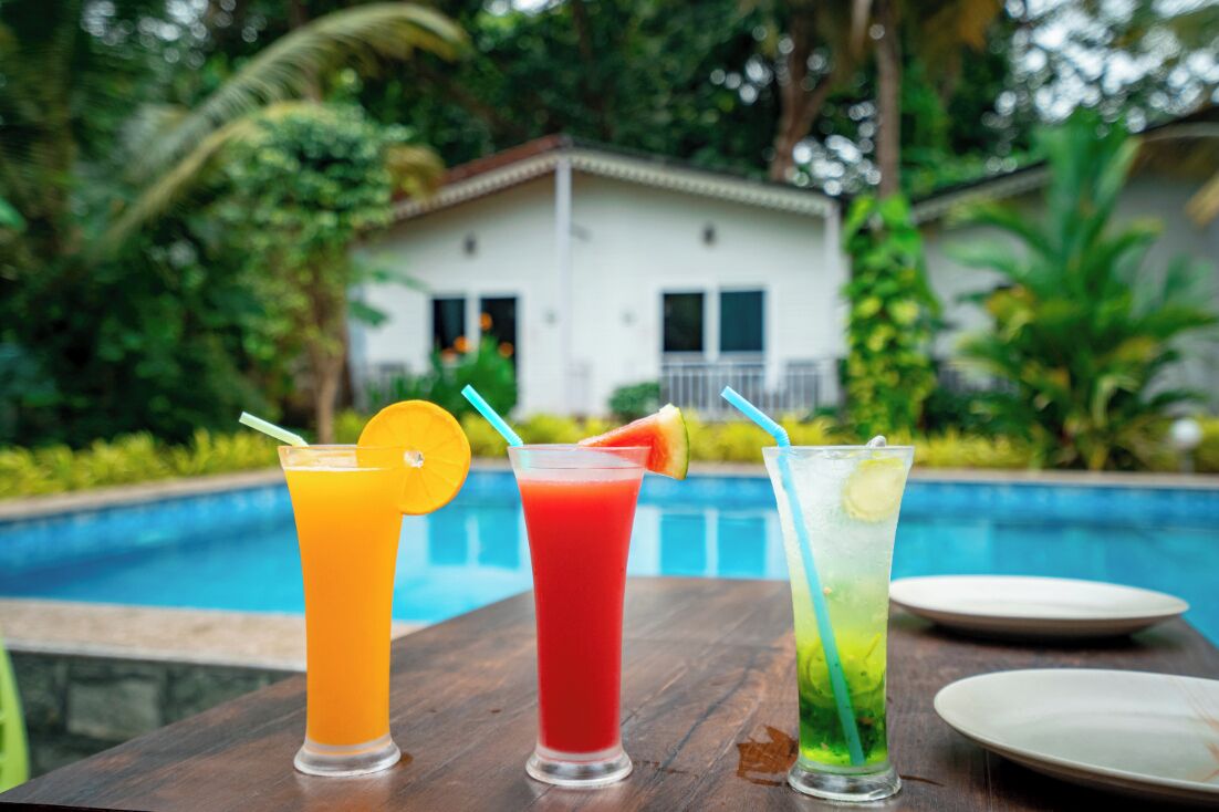 Three drinks on a table by a pool with a house in the background.