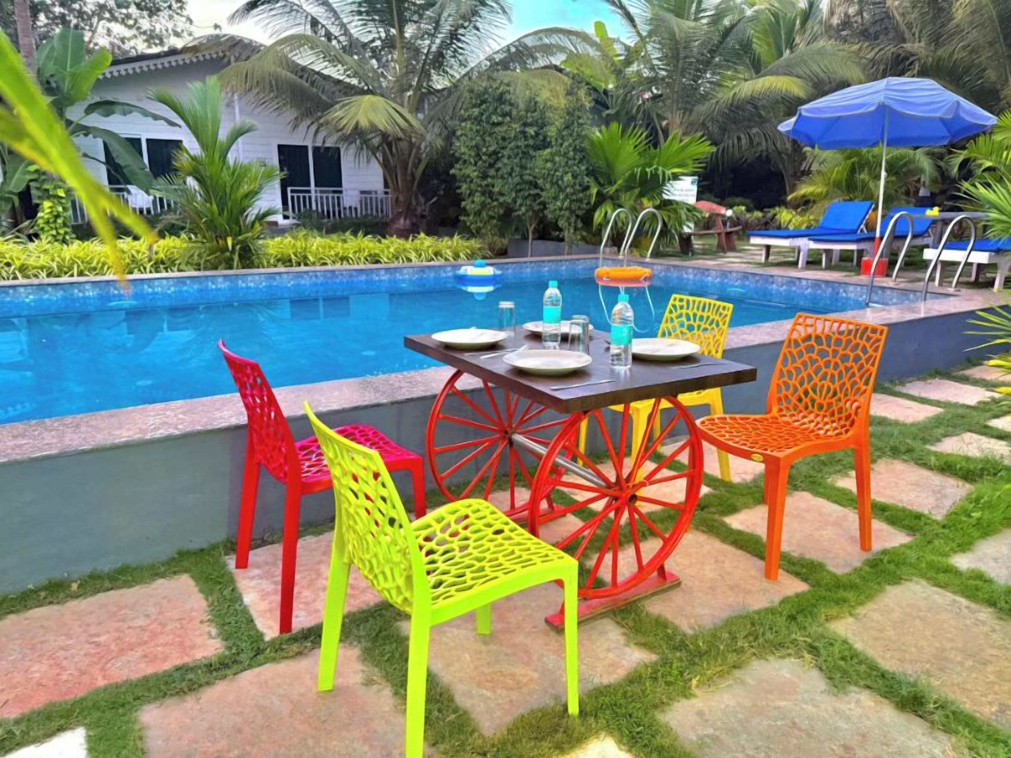 A poolside area with a table set for two, surrounded by colorful chairs, and a blue umbrella.