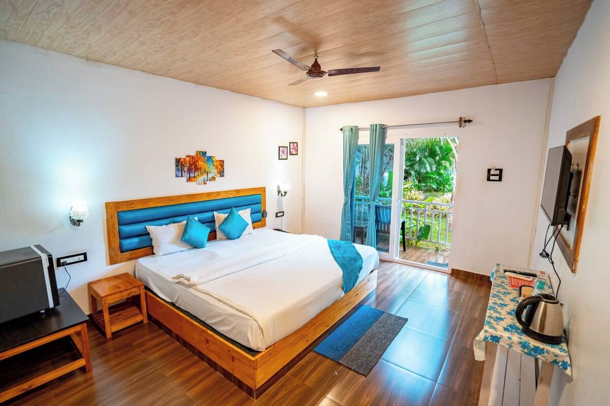 A bedroom with a wooden ceiling, a bed with blue and white bedding, a small bedside table, a TV, and a balcony with a view of greenery.