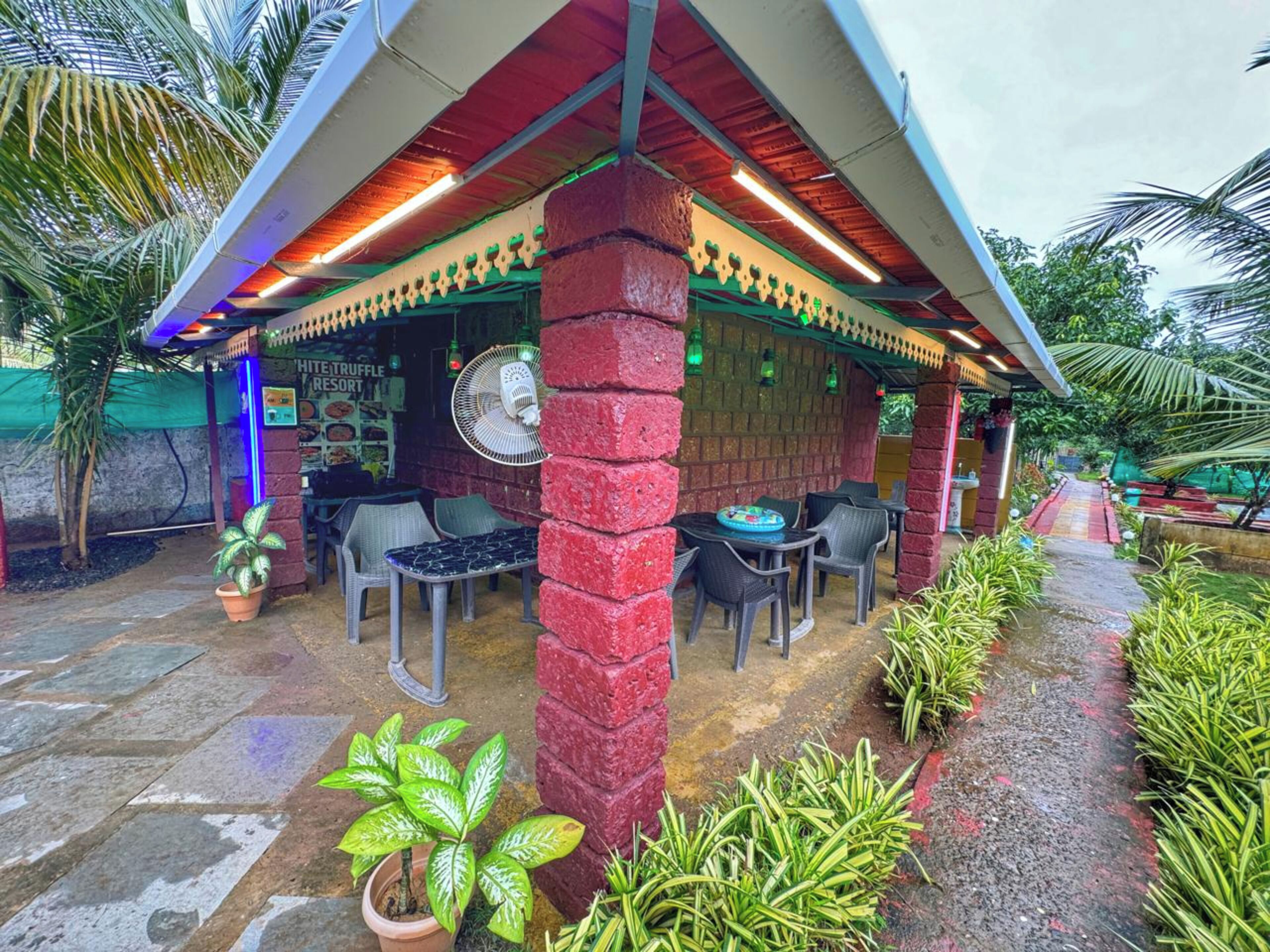 A covered outdoor seating area with brick pillars, tables, and chairs, surrounded by potted plants and palm trees.