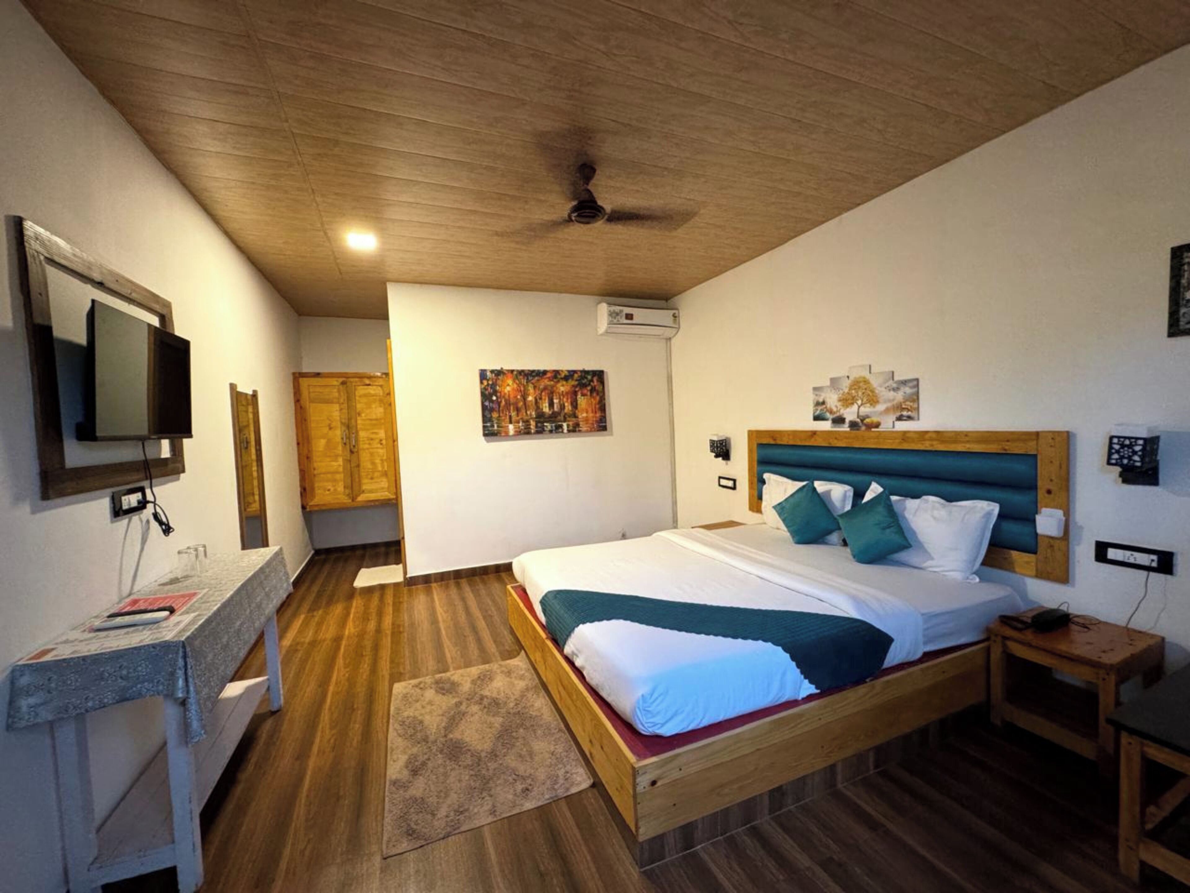 Quarto com teto de madeira, cama com roupa de cama verde-água e branca, televisão na parede e um quadro sobre a cama.