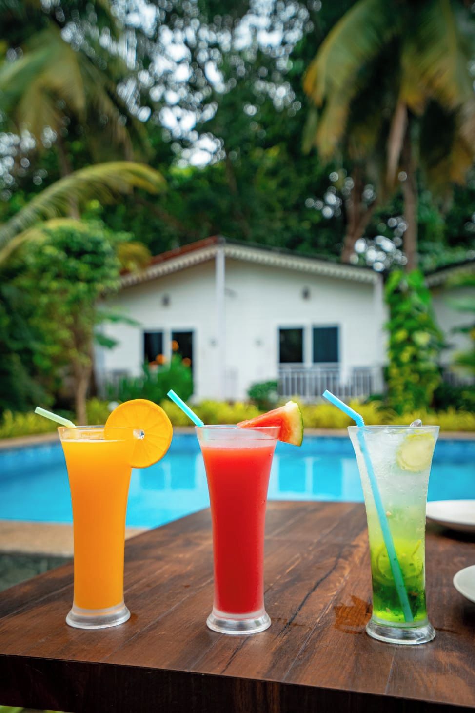 Three drinks on a wooden table with a pool and a house in the background.