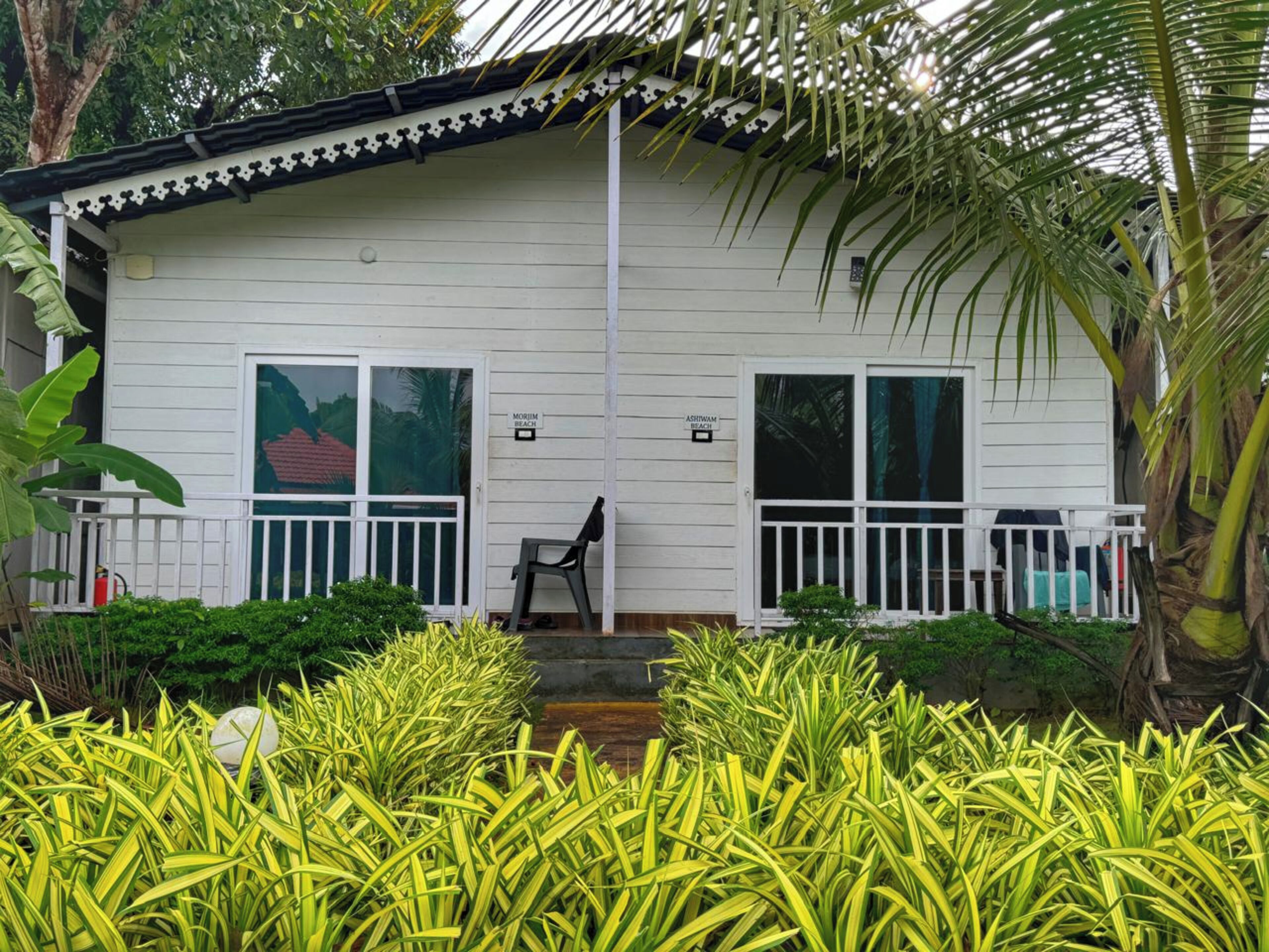 A white two-story house with a balcony and a covered porch, surrounded by lush greenery.
