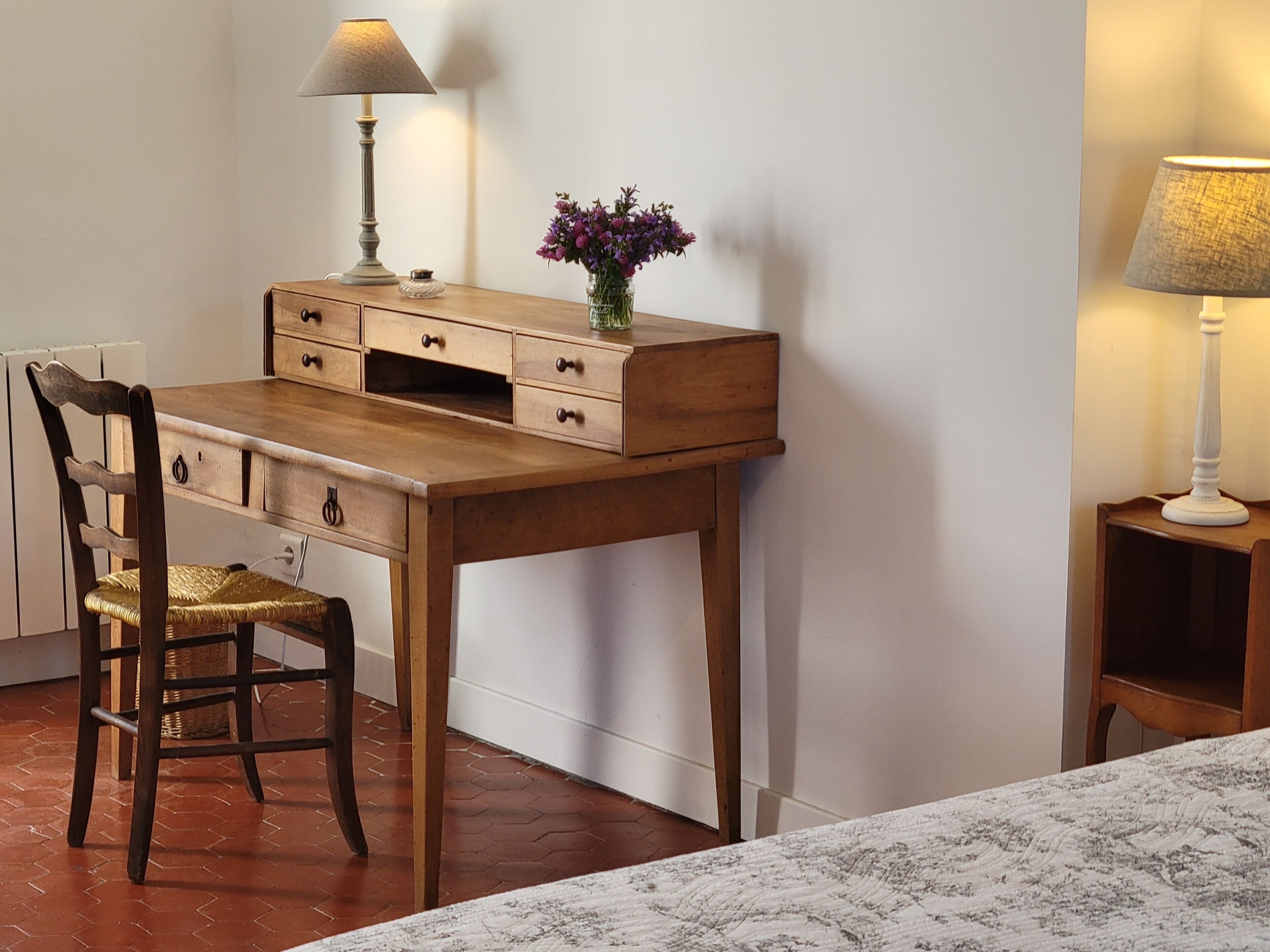 Een houten bureau met lades, een stoel, een lamp en een vaas met bloemen.