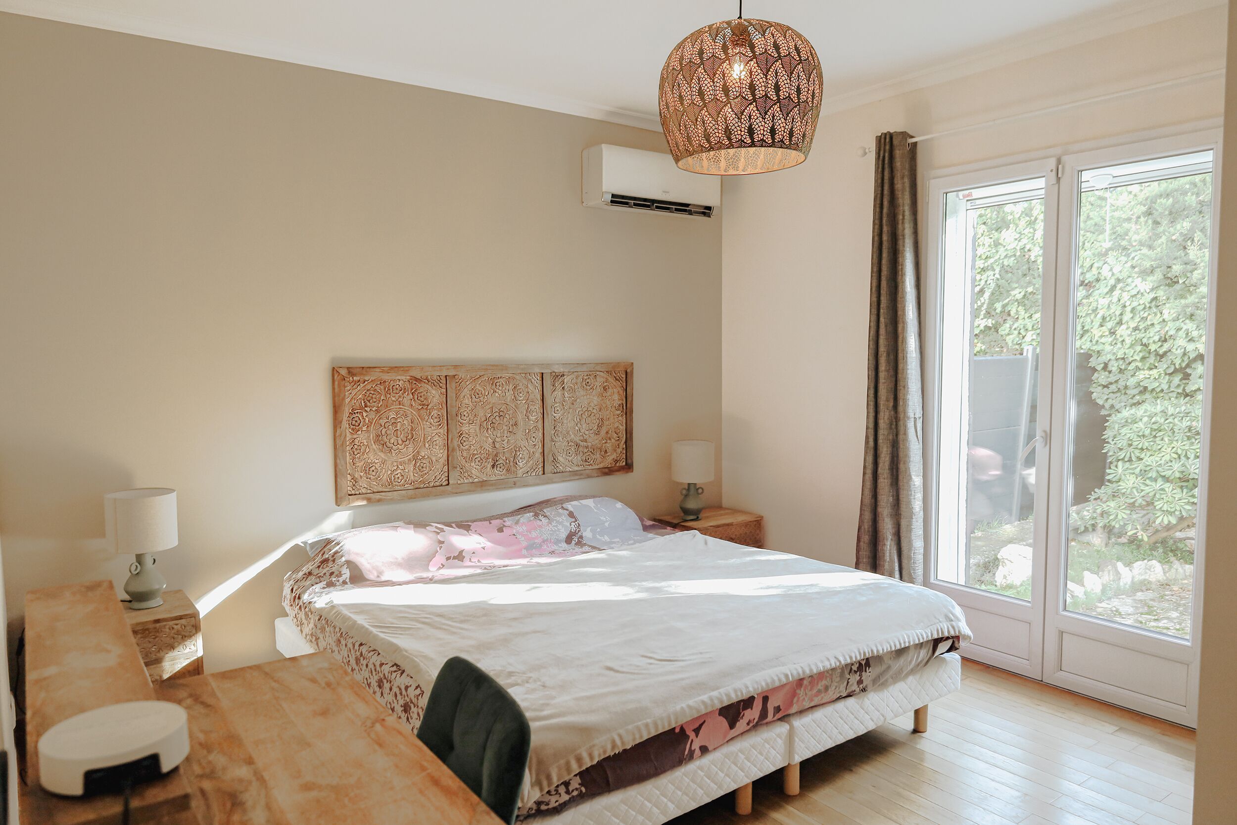 A bedroom with a large bed, bedside tables, a desk, and a window with a view of greenery.