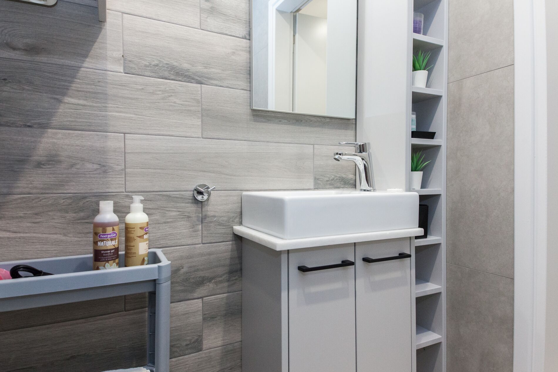 Un baño moderno con baldosas de madera gris, un lavamanos blanco y un espejo encima. Hay dos botellas en un estante y una planta en un estante a la derecha.