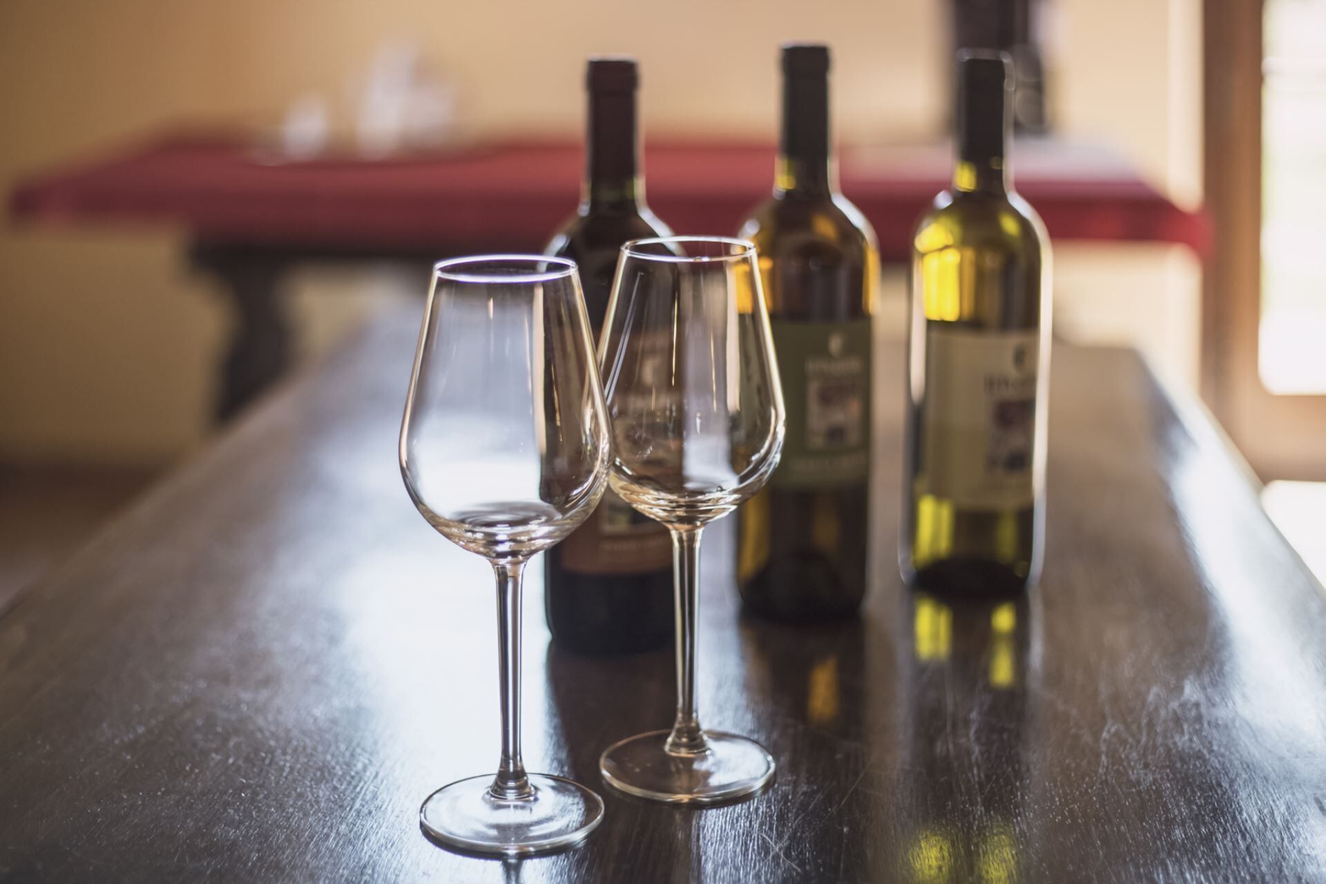Dos copas de vino vacías sobre una mesa de madera, con tres botellas de vino al fondo.