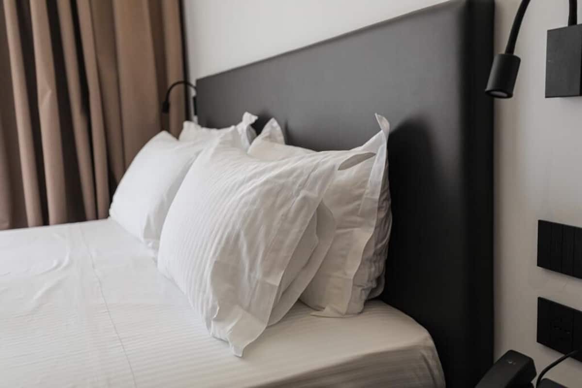 Un lit bien fait, avec du linge de lit blanc et des oreillers, une tête de lit sombre et un luminaire fixé au mur.