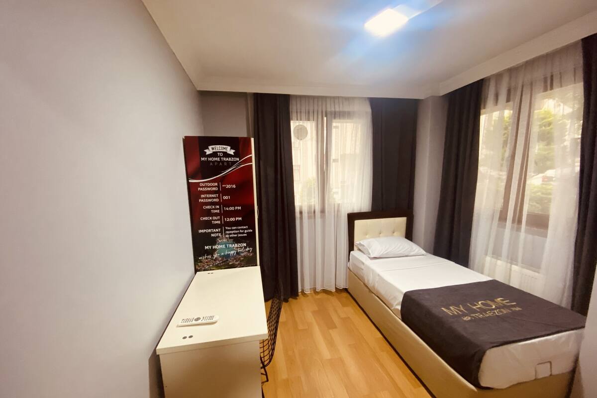 Um quarto com uma cama, uma escrivaninha e uma janela com cortinas. Há um letreiro de boas-vindas na parede.