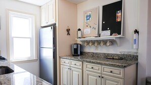 A kitchen with white cabinets, a granite countertop, a refrigerator, a coffee maker, and a wall-mounted shelf with mugs.