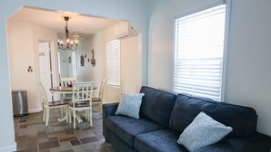 A living room with a dark blue sofa, two patterned pillows, a chandelier, a dining area with a white table and chairs, and a large window with blinds.