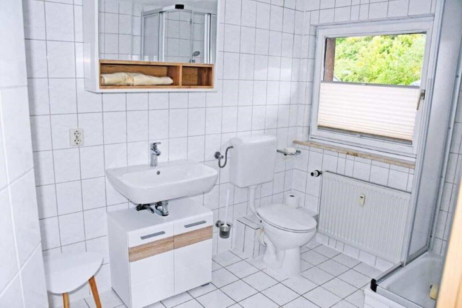 A bathroom with a white sink, toilet, and shower. There is a window with a view of greenery outside.