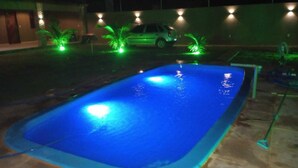 A swimming pool with a blue illuminated edge, surrounded by palm trees and a parked car. - Furnished and cozy beach house in Trairí - CE, close to beautiful beaches! (Trairi)