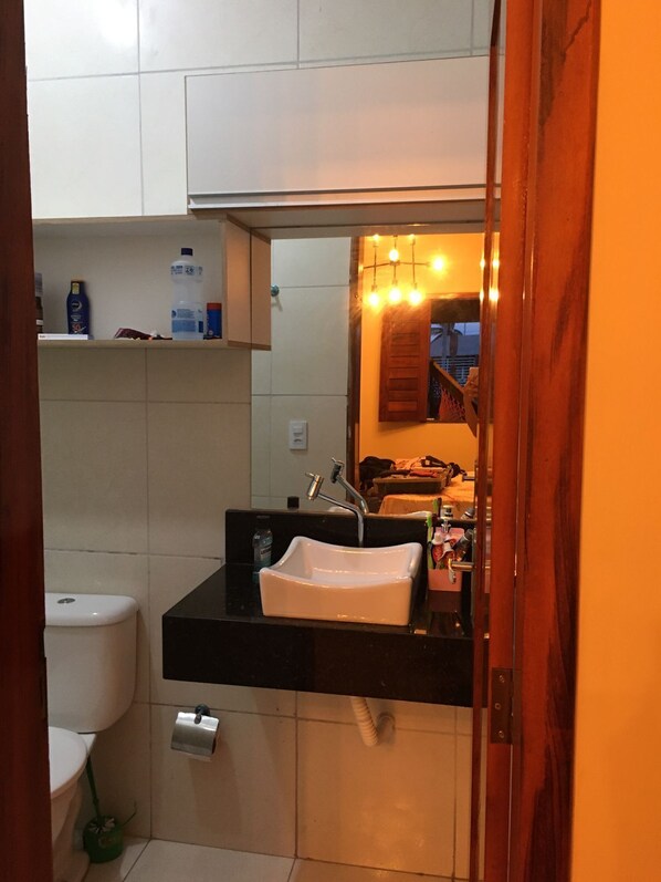 Un baño pequeño con un lavamanos blanco, encimera negra y un espejo que refleja una habitación con cama y mesita de noche.
