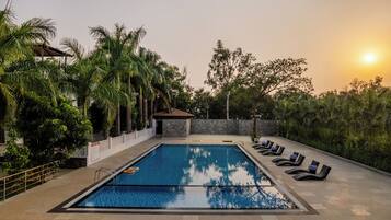 Una piscina rectangular con un borde de baldosas, rodeada de palmeras y un muro de piedra. Hay sillones de descanso a lo largo de la piscina.